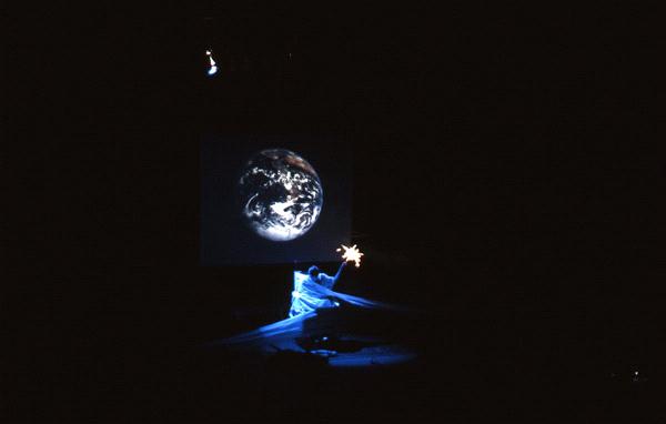 A dimly lit scene with a projection of Earth against a dark background. A person below the projection operates equipment and holds a sparkler, creating a subtle glow and contrast in the darkness.