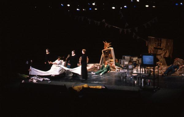 A theatrical stage with actors in dramatic poses. The scene includes a bed, scattered sheets, and various props like televisions and a statue. Dim lighting and a dark background create a mysterious atmosphere.