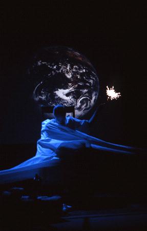 A person draped in white fabric sits on a dark stage, reaching out with one hand towards a small, glowing light. Behind them is a large image of Earth. The scene is illuminated by blue lighting, creating a dramatic and ethereal atmosphere.