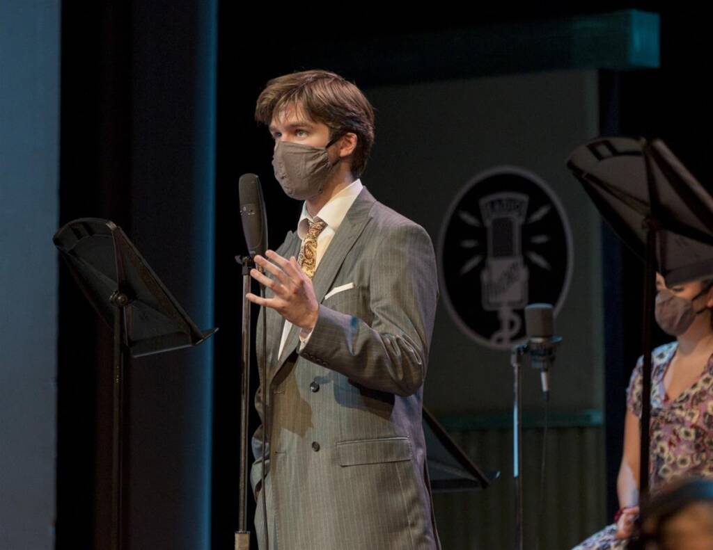 A person in a suit and tie speaks into a microphone on stage. They are wearing a mask and are gesturing with one hand. Another person, also masked, is visible in the background near a music stand. Stage lights and equipment are partially visible.