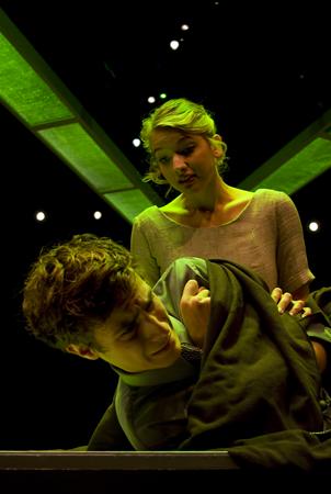 A young woman stands under a green light, looking down with concern at a kneeling man who appears distressed, clutching a dark blanket. The scene is set against a dark background with small white lights.