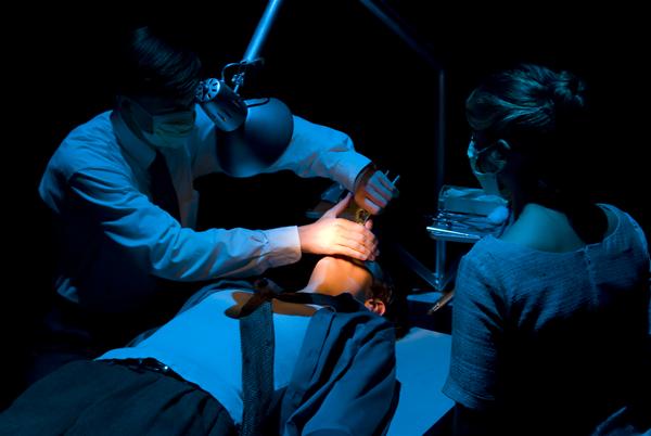 A dentist in a well-lit, dark room examines a patient lying on a chair. The dentist wears a mask and glasses, while an assistant stands nearby, observing the procedure. Shadows and focused lighting create a dramatic atmosphere.