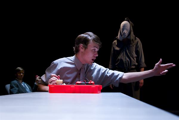 A man seated at a table passionately gestures with his right hand. In front of him is a red device with wires. In the background, a hooded figure stands, face obscured. Another person sits to the left, partially visible. The scene is dimly lit.