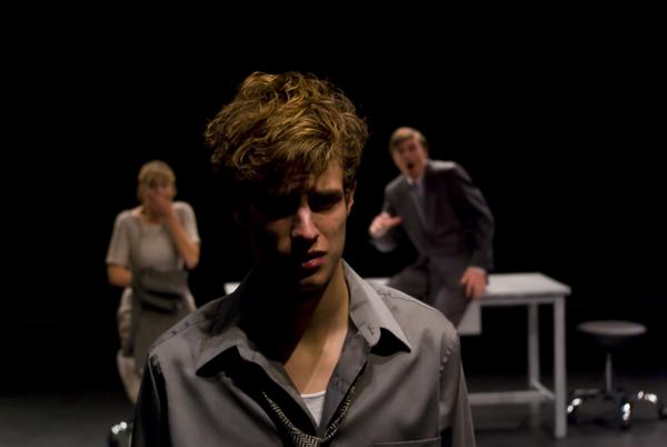 A young man with curly hair stands in the foreground, looking distressed. In the background, a woman and a man in a suit are at a desk, both appearing shocked. The scene is set on a dark stage.