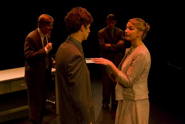 A woman in a suit gesturing toward a man standing in front of her, while two men in suits observe in the background. They appear to be on a dimly lit stage or set, with a table nearby.