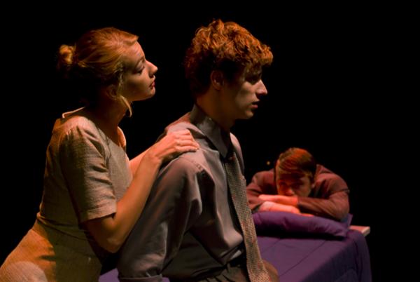 Three people in a dimly lit scene: a woman with her hand on a sitting man's shoulder, both looking forward, onstage. A man in a suit is lying on a bed in the background, resting his head on his arms, eyes closed.