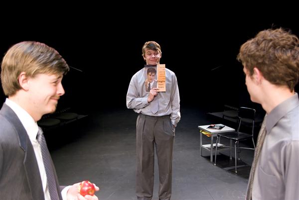 Three men in formal attire are on a stage. The man in the center holds a book with a photo on the cover. The man on the left holds an apple, and the man on the right observes. A few chairs are in the background.