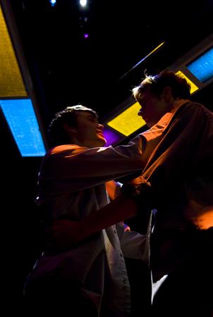 Two people in dim lighting face each other closely, creating a dramatic silhouette. They are surrounded by colorful, stained glass-like lights in shades of blue, yellow, and purple, set against a dark background.