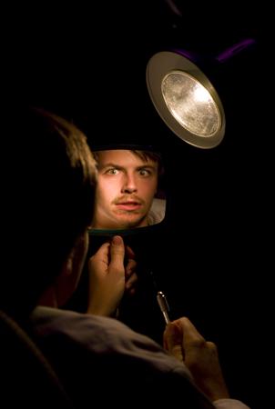 A person looks into a hand-held mirror, reflecting a surprised expression. The scene is dimly lit with a focused spotlight illuminating the mirror and the person's face. The background is dark.