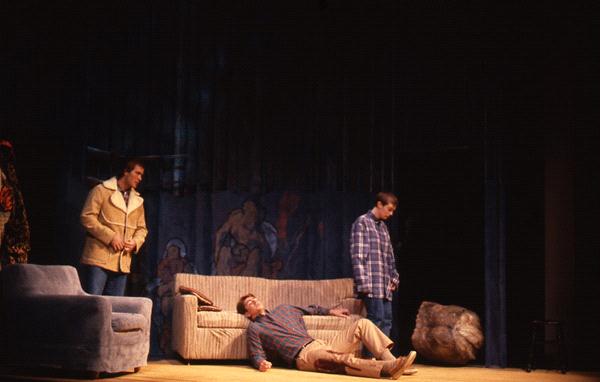 Three people are on stage in a dim theater setting. One person lies sprawled on a sofa, while two others, wearing jackets, stand nearby. Behind them is a backdrop with artwork. A chair and some props are visible on stage.