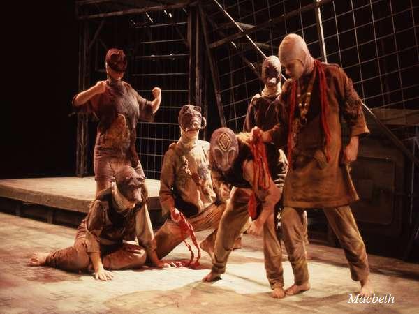 A theatrical scene depicting six masked actors in ragged clothing. They are engaged in a dramatic pose on a dimly lit stage with a metal grid backdrop. The scene conveys intensity and action.