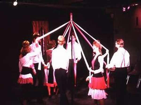 A group of people in traditional attire are dancing around a maypole, holding colorful ribbons. The setting appears to be indoors, with dim lighting highlighting the dancers.