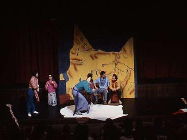 Actors perform on a stage in front of a painted backdrop. Some are standing, while others are sitting on a bench. The scene likely depicts a dramatic or comedic performance in a theater setting.