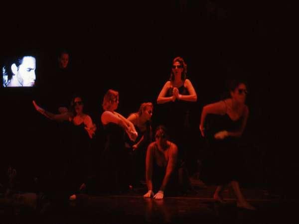 A group of performers in dark clothing pose on a dimly lit stage, striking various dramatic positions. A small screen in the background displays a person's face. The atmosphere is theatrical and intense.