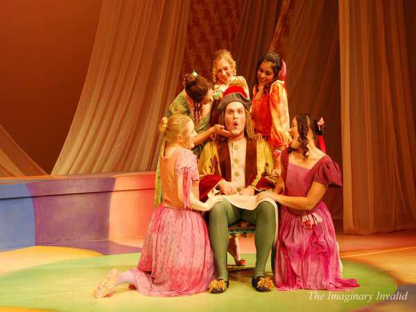 A theatrical scene from "The Imaginary Invalid" shows a man in colorful period costume seated center stage, surrounded by six women in vibrant dresses. They gesture animatedly, creating a lively atmosphere against a draped fabric backdrop.