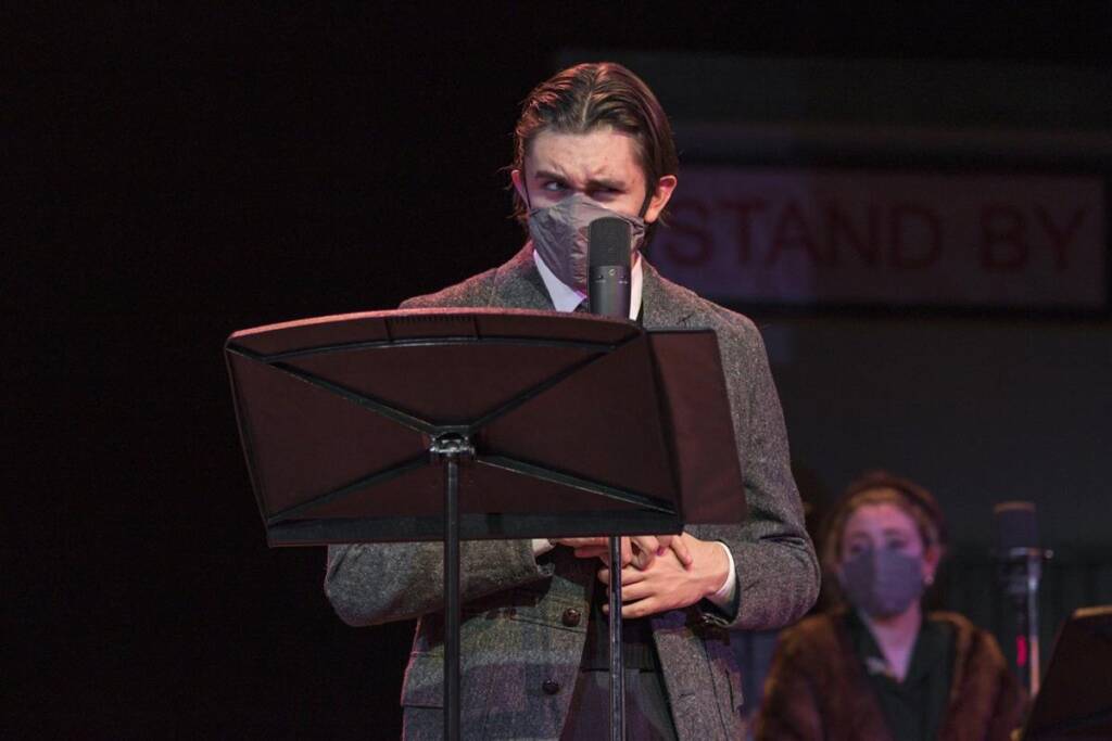 A person wearing a tweed coat and a face mask stands at a microphone, reading from a music stand. Another person, also masked, is in the background. The setting appears to be a performance or recording space.