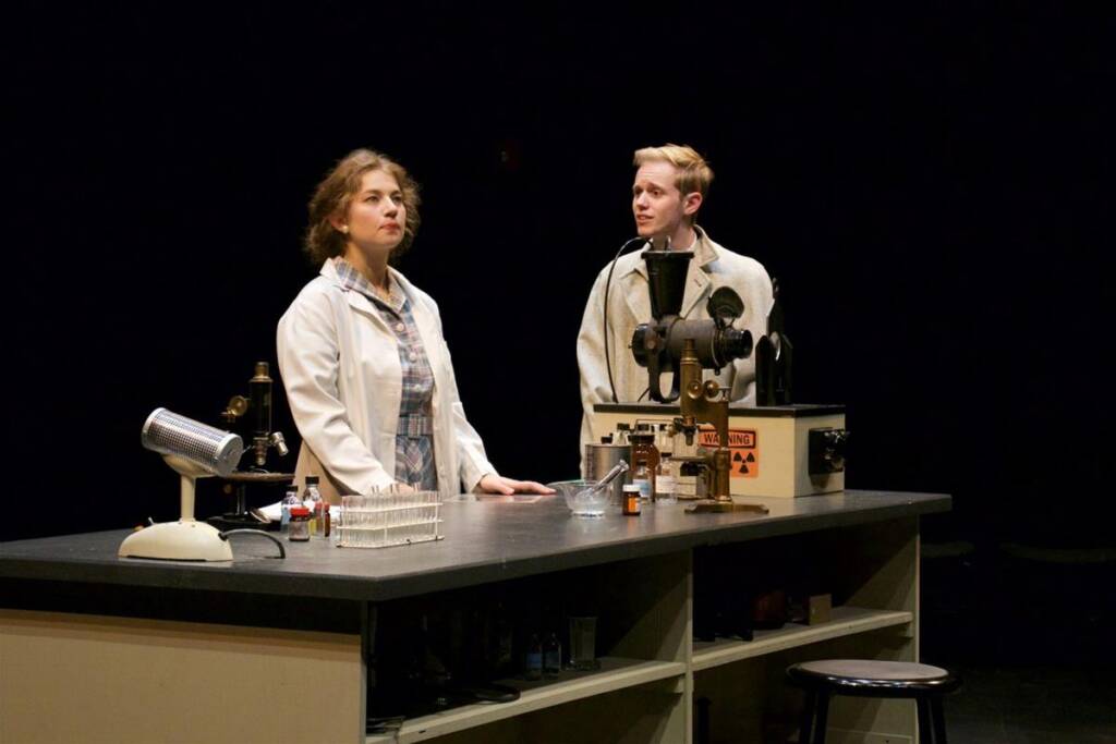 Two people in lab coats stand behind a table filled with scientific equipment in a dimly lit room. The table holds test tubes, a microscope, and other lab instruments. The scene suggests a laboratory or scientific setting.