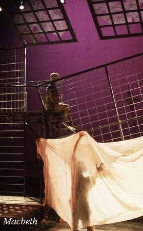 A theatrical stage with a modern set design depicting a scene from "Macbeth." A person sits on an elevated structure with grid patterns, while another holds a large, translucent fabric below, creating dramatic shadows. Lighting has a purple hue.