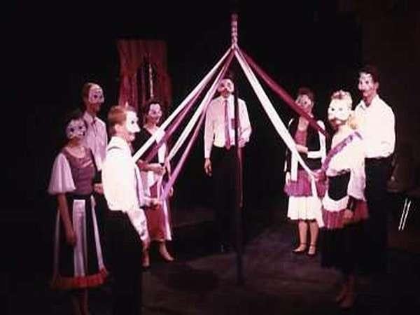 People in masks and costumes dance around a maypole with ribbons on a dimly lit stage.