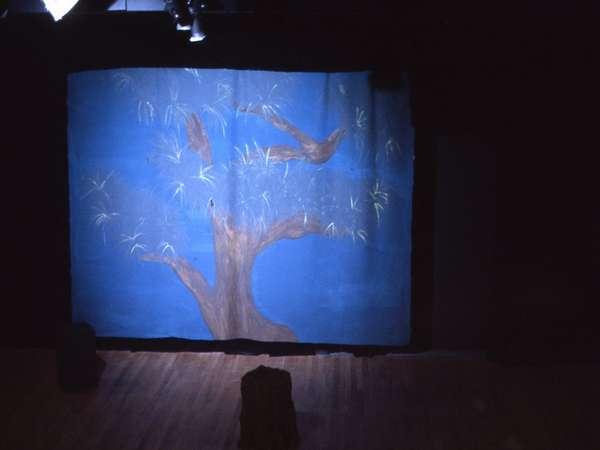 A dimly lit stage features a painted backdrop of a large tree with bare branches against a blue background. The wooden floor and a solitary dark object in the foreground are visible.