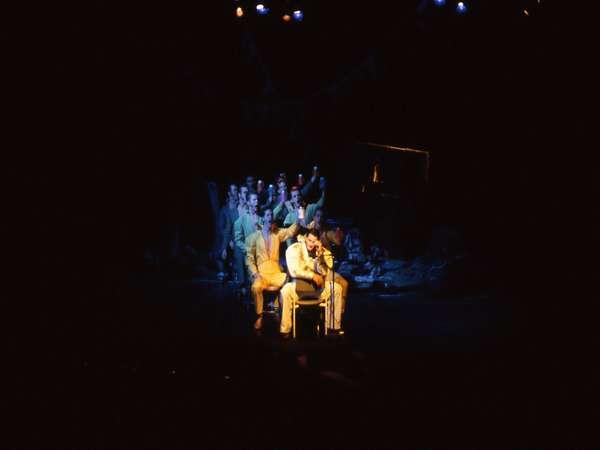 A person sits on a chair on a dimly lit stage, surrounded by a group of people standing closely behind. The background is dark, with minimal lighting highlighting the figures. The scene appears theatrical and dramatic.