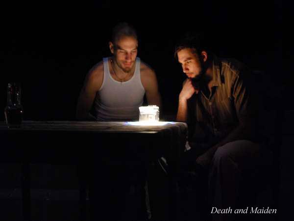 Two people sit at a table in dim light, focused on a small, lit birthday cake. The background is dark, highlighting the cake's glow. The words "Death and Maiden" are faintly visible in the bottom right corner.