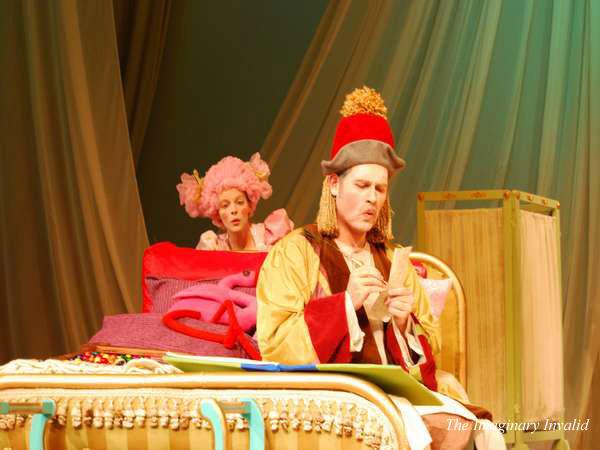 Two performers in ornate costumes sit on stage. One, wearing a red hat and gold robe, appears focused on an object in their hands. The other, in a pink wig and elaborate dress, looks on. The backdrop features draped fabric.