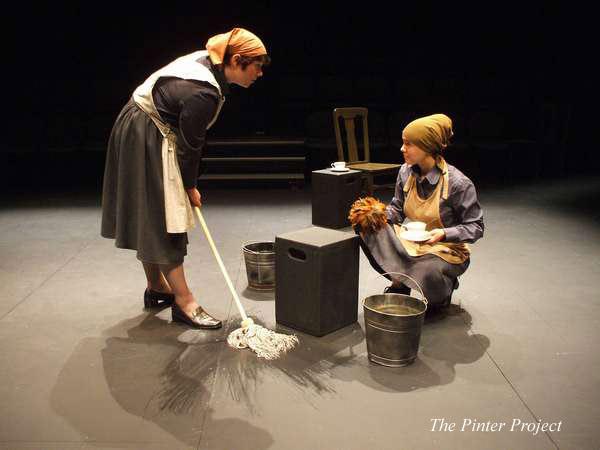 Two people in costumes resembling domestic workers are on stage. One is standing and mopping, while the other is sitting, holding a rag and a brush. They are surrounded by buckets. The setting is dimly lit, offering a theatrical ambiance.