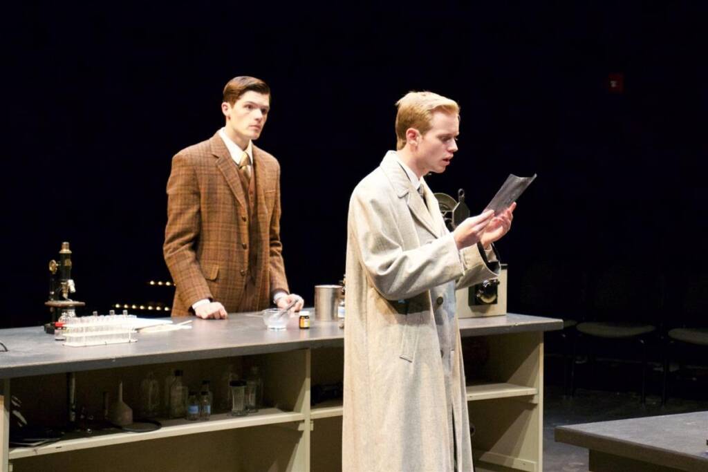 Two men in a lab setting, one in a plaid suit stands by the bench with lab equipment, while the other in a light trench coat examines a document. The background is dark, focusing on their solemn expressions.