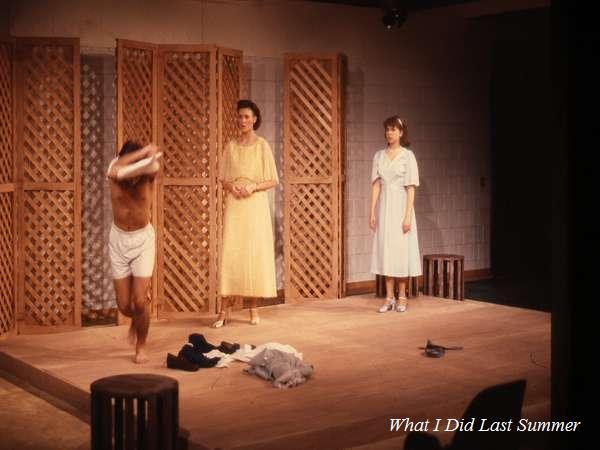 A stage performance featuring three actors. One person is in motion on the left, wearing shorts and swinging an arm, while two others stand watching; one in a yellow dress and the other in a white dress. The set has wooden lattice panels.