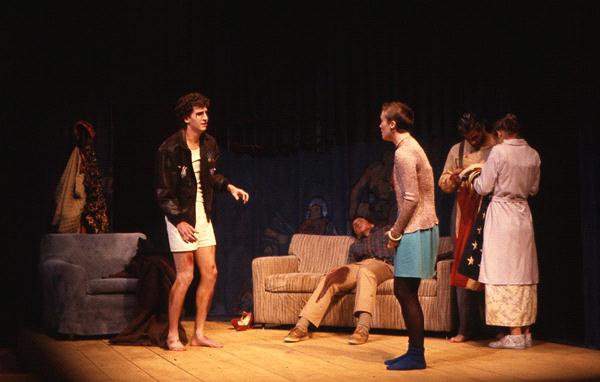 A stage scene with five actors. One actor stands in a coat and boxers, gesturing. Another actor faces them, wearing a skirt. Three actors are on a couch; one is seated and appears asleep, while the others stand nearby. A blue chair is visible on stage.