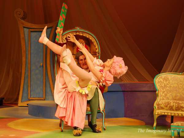 A performer in a pink dress is playfully sitting on another performer's lap, holding their own leg up high. Both are in colorful costumes, with an elaborate set featuring whimsical furniture in the background.