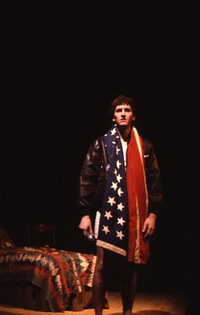 A person stands on a dimly lit stage, wearing a dark jacket and draped in an American flag. A bed with a similar flag-themed blanket is in the background. The scene conveys a dramatic or theatrical setting.