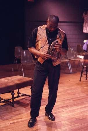 A man stands on a wooden stage holding an instrument, possibly a saxophone. He is wearing a patterned vest over a black shirt and pants. Empty chairs are visible in the background.