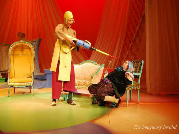 Two actors are on a colorful stage. The one on the left, wearing a yellow hat and coat, uses an oversized syringe on the other actor, who is wearing colorful clothing and sitting on a chair. The set has vibrant colors and vintage furniture.