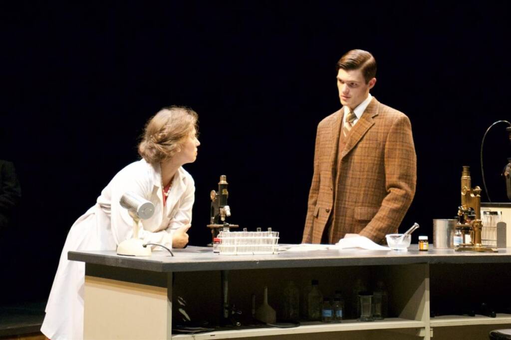 Two people stand at a laboratory table with scientific equipment, including microscopes. The person on the left wears a lab coat and leans forward, while the person on the right is in a brown suit, looking thoughtfully at the other.