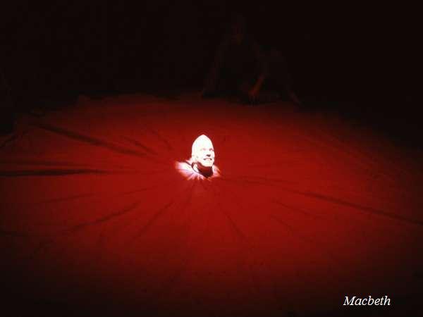 A person with a light shining on their face emerges from the center of a large, dark red circular fabric. The rest of the area is in shadow. The word "Macbeth" is in the bottom right corner.