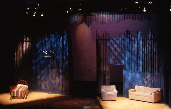 The image shows a theater stage set with a bed on the left, a single chair in the center, and a sofa on the right. The backdrop features abstract art with lighting creating striped shadows across the scene.