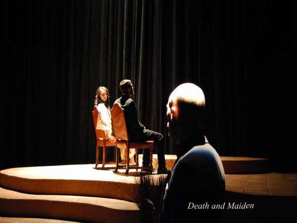 Two individuals sit on chairs facing each other on a dimly lit stage with a third person standing in the foreground. The scene has a dramatic atmosphere, with the words "Death and Maiden" visible at the bottom.