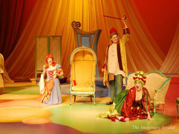 Three actors in colorful period costumes perform dramatically on stage under warm lighting. One stands with a raised cane, while the others kneel and gesture expressively. An ornate chair and draped backdrop are visible. Text says "The Imaginary Invalid.