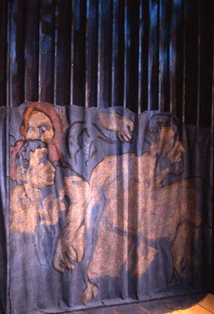 A textured mural on a vertical corrugated surface depicts abstract human figures intertwined. The figures are painted in earthy tones, including brown and orange, against a dark background with blue lighting from above.