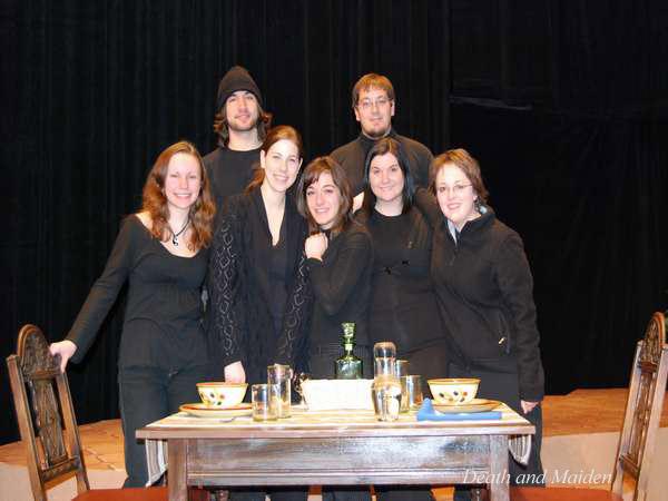 Seven people wearing black clothing stand behind a table set with dishes, glasses, and a bottle. They are on a stage with a dark curtain backdrop. Two wooden chairs are partially visible on either side of the table.