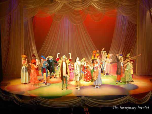 A theatrical performance featuring a group of actors in colorful costumes on stage. The set includes draped fabric and warm lighting. The scene is dynamic, with performers in animated poses. The title "The Imaginary Invalid" is visible.