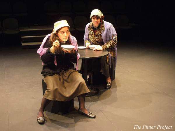 Two actors in vintage clothing sit at a small round table on stage, holding bowls. One, in the foreground, appears to be eating. The stage is dimly lit with empty seats in the background. "The Pinter Project" is written in the corner.