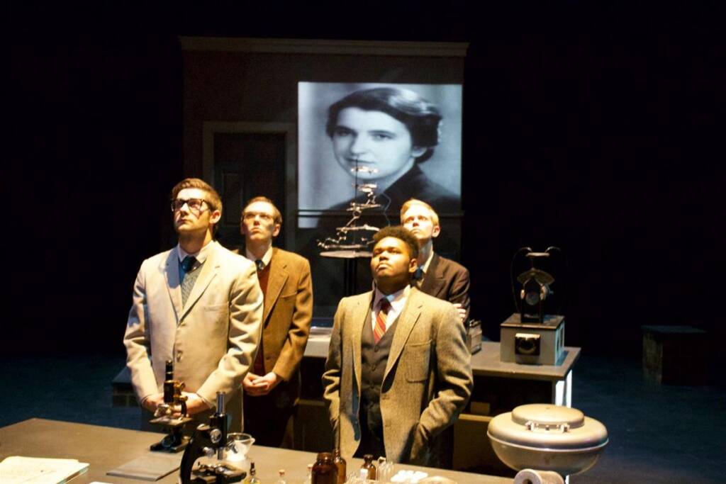 Four men in suits stand facing forward in a dimly lit room with laboratory equipment around them. A black-and-white portrait of a woman is projected in the background.