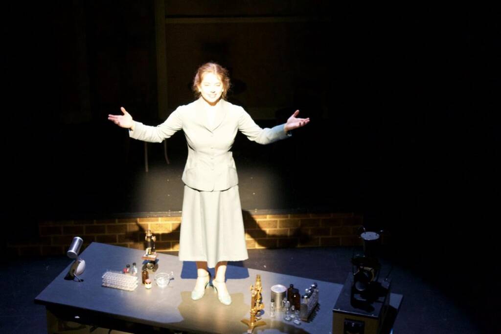 A woman stands on a dimly lit stage with her arms outstretched. She is wearing a light-colored suit and shoes. The table in front of her holds scientific equipment, and a spotlight shines on her, emphasizing her presence.