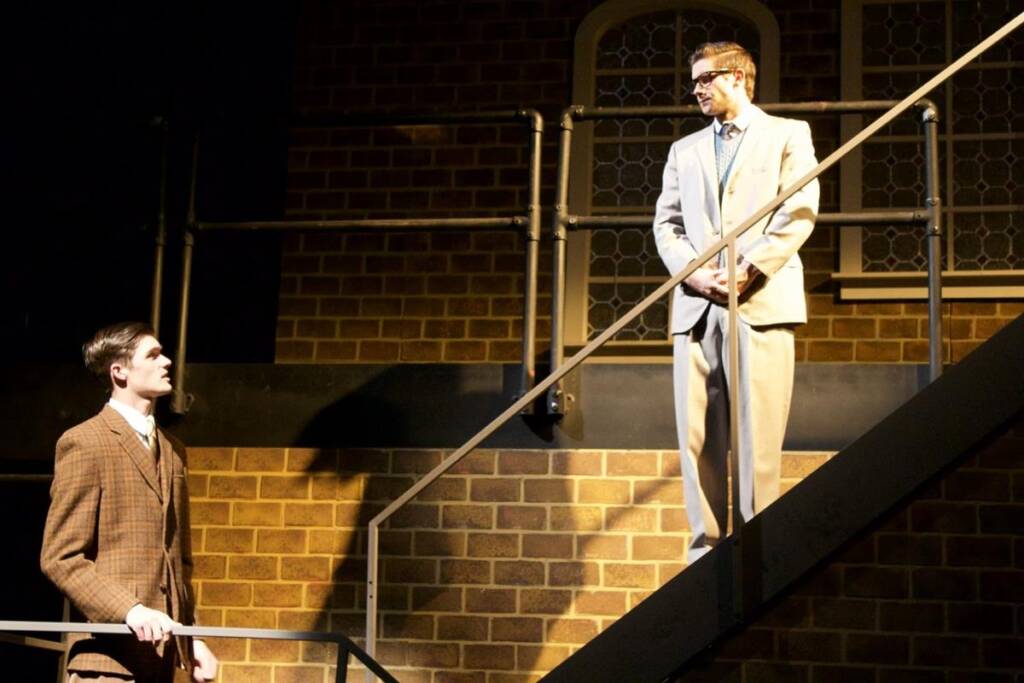 Two men in suits stand on a staircase on a stage. One man is higher up, wearing glasses and a beige suit. The other man is lower, wearing a brown suit. They are set against a brick wall background with metal railings.