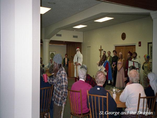 A group of people in a room with several seated at tables and others standing. Some are in costumes, suggesting a theatrical performance or event. The text "St. George and the Dragon" is visible in the corner.