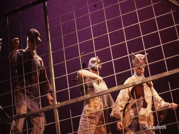 Three masked performers in costume stand behind a metal grid on stage, under dramatic lighting. The setting appears to be theatrical, with a purple background. The word "Macbeth" is visible in the bottom right corner.