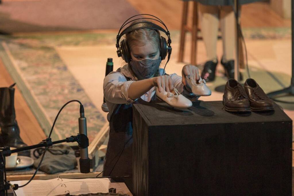 A person wearing headphones and a mask is seated, holding a pair of small tap shoes above a black box. Nearby, an adult pair of shoes is placed on the box. Microphones and a notepad are visible in the setup.